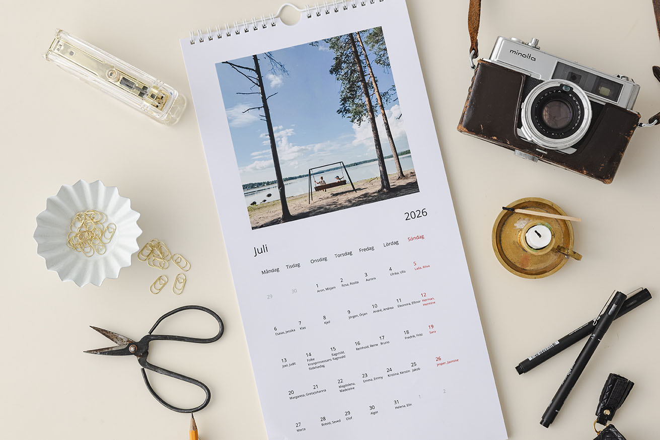 Fotokalender uppslagen med juli måndad 2026, placerad på ett borde omgivet av detaljer som en kamera och kontorsmaterial.